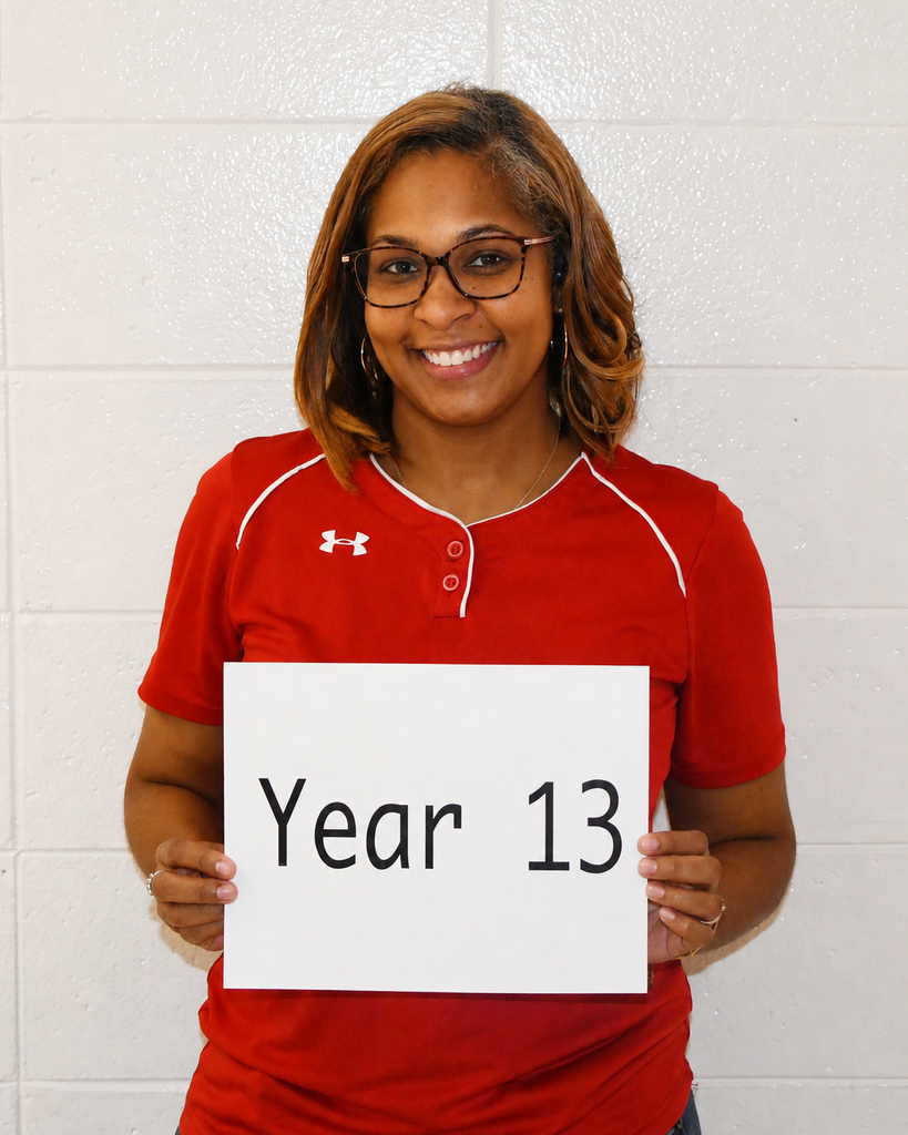 A woman in a red polo shirt holds a white sign with the text "Year 13" against a white wall.