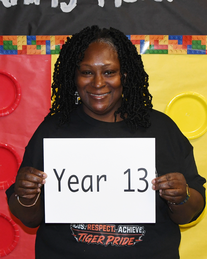Woman smiling, holding a white sign that reads "Year 13" in front of a colorful backdrop with Lego blocks.