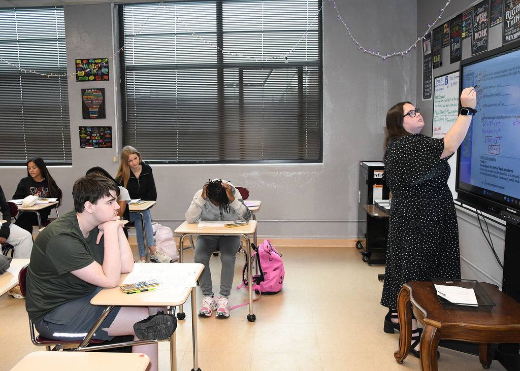 A classroom with students seated at desks. A woman in a black dress stands by a whiteboard.