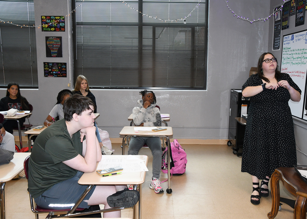 A classroom with students seated at desks. A woman in a black dress stands near a whiteboard, speaking to the class.