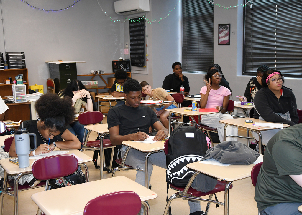 A classroom with desks, students seated, and a person leaning on a desk. An air conditioner, shelves, and windows are visible.