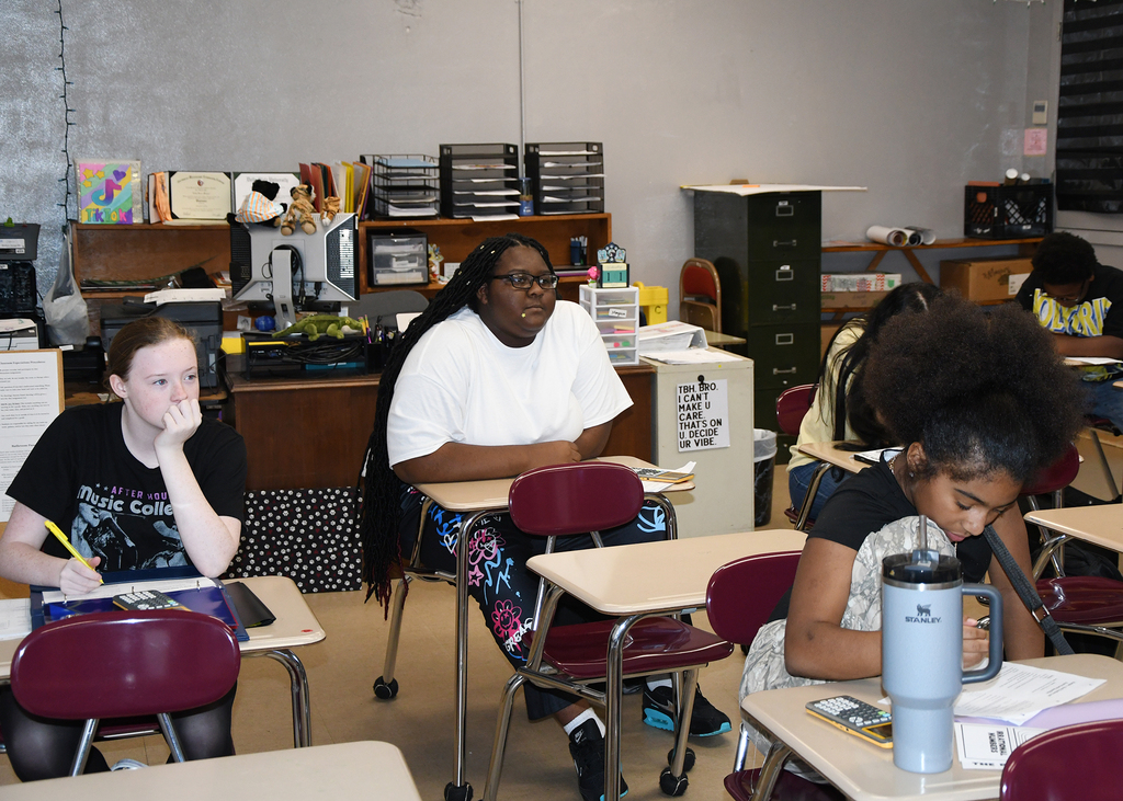 A classroom with several students seated at desks, engaged in writing or reading activities. The desks are arranged in rows, and various items are on the desks.
