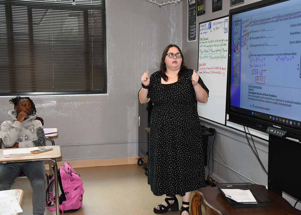 Woman in black dress stands in front of a classroom, explaining something with hand gestures.