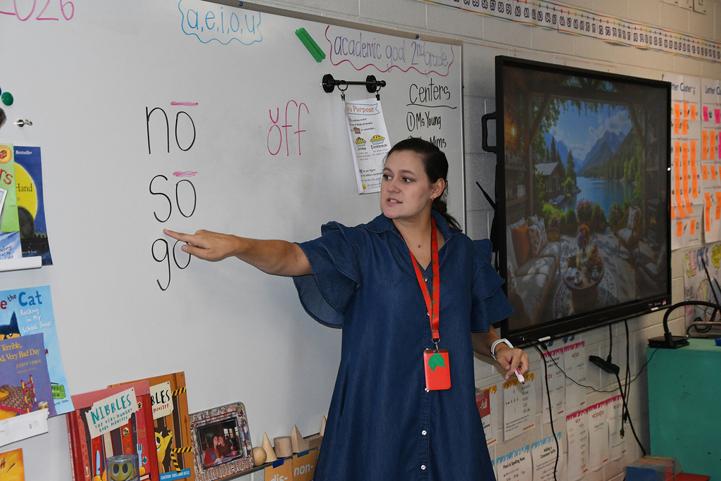 A teacher points to a whiteboard with text while a monitor displays an image, in a classroom with books and materials.