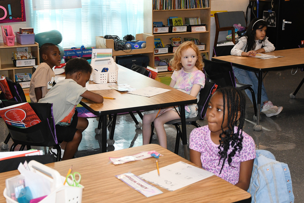 Children sitting at desks in a classroom, engaged in activities with papers and pencils. Background features shelves with books.