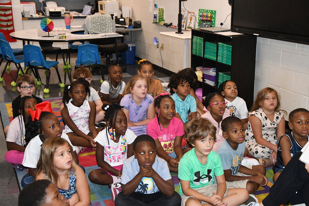 Classroom with children seated on the floor. Some children are wearing glasses, sneakers, and bracelets.