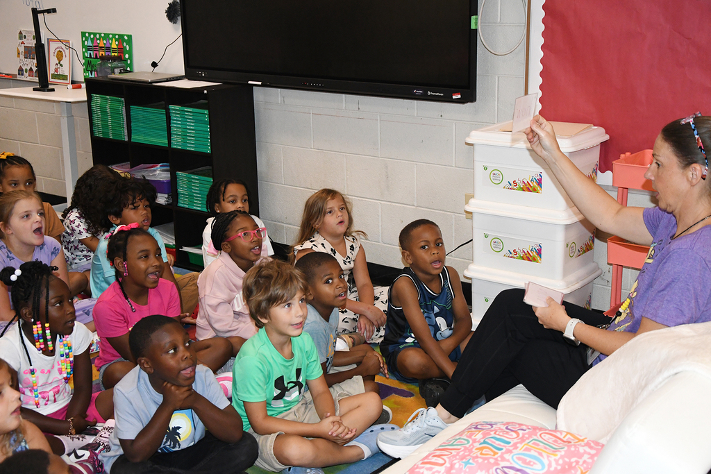 Group of children seated around a woman holding a book, in a classroom with a television and shelves.
