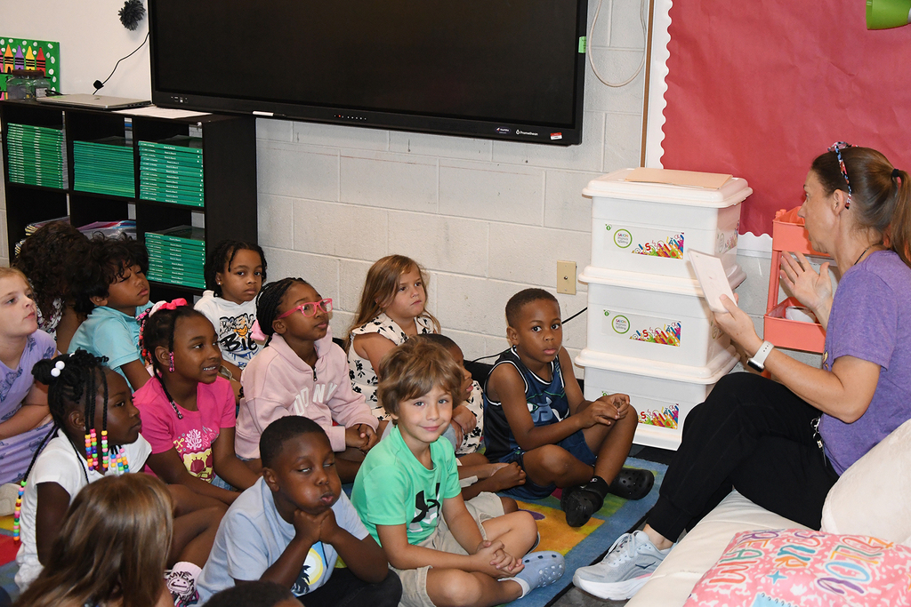 A group of children in a classroom listening to a woman read a book.