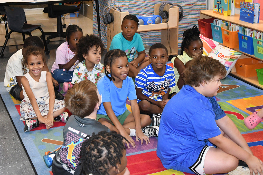 A group of children and a teacher sit on a colorful floor mat in a classroom.