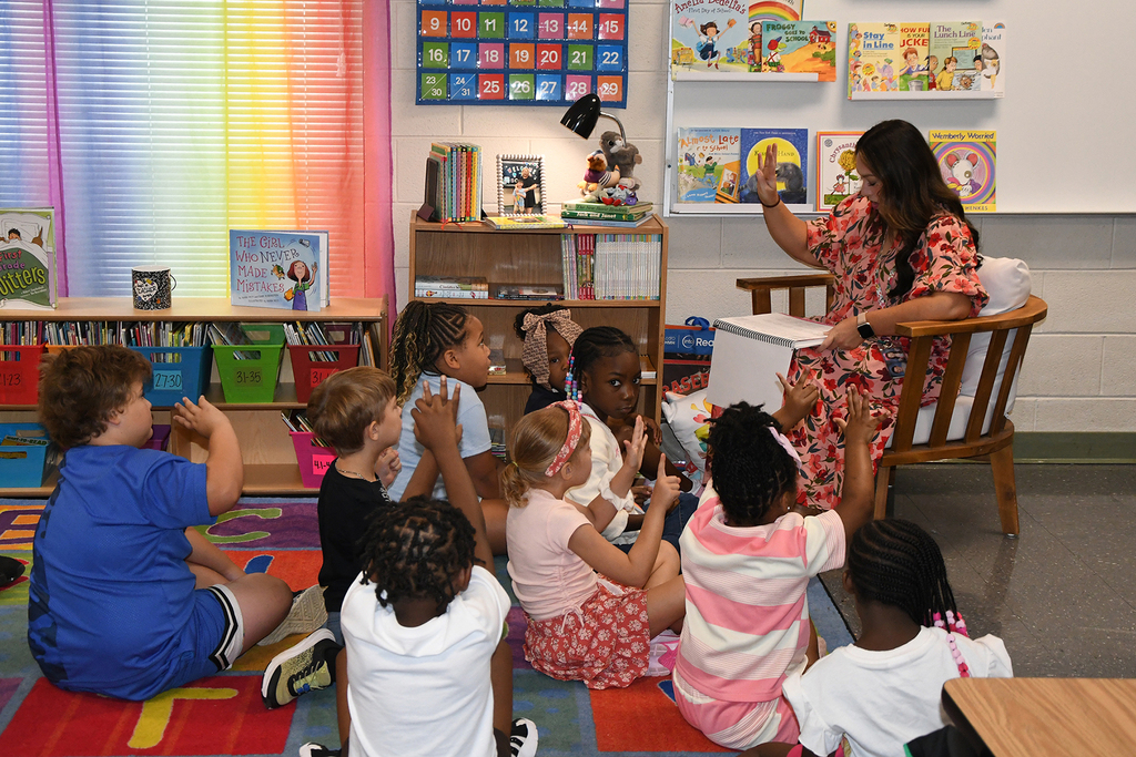 A woman reads a book to children sitting in a circle in a classroom with shelves of books.
