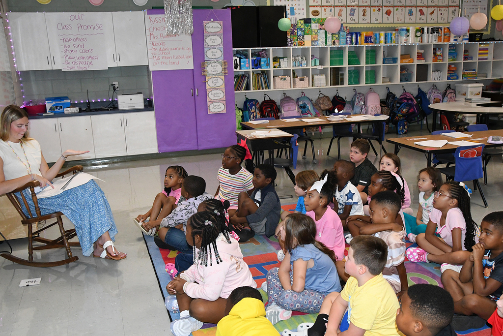 A teacher sits on a rocking chair, instructing a group of children sitting on a rug in a classroom.