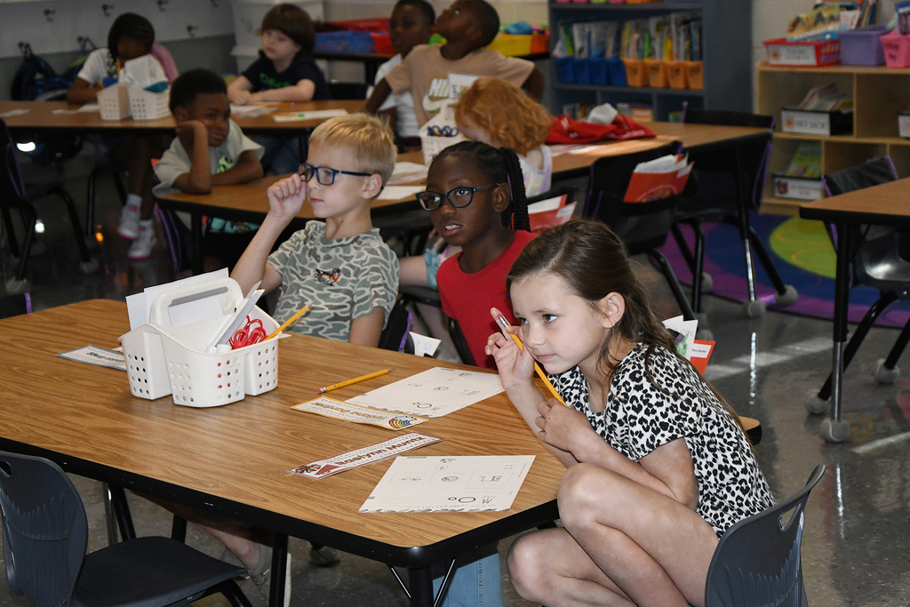 Students sit at desks, using pencils to write on paper. A girl in the front has a pencil in her mouth.