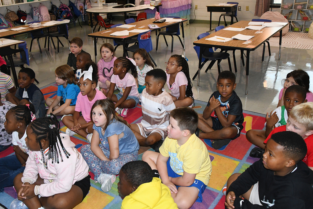 Group of children sit cross-legged on a colorful carpet, facing a teacher in a classroom.