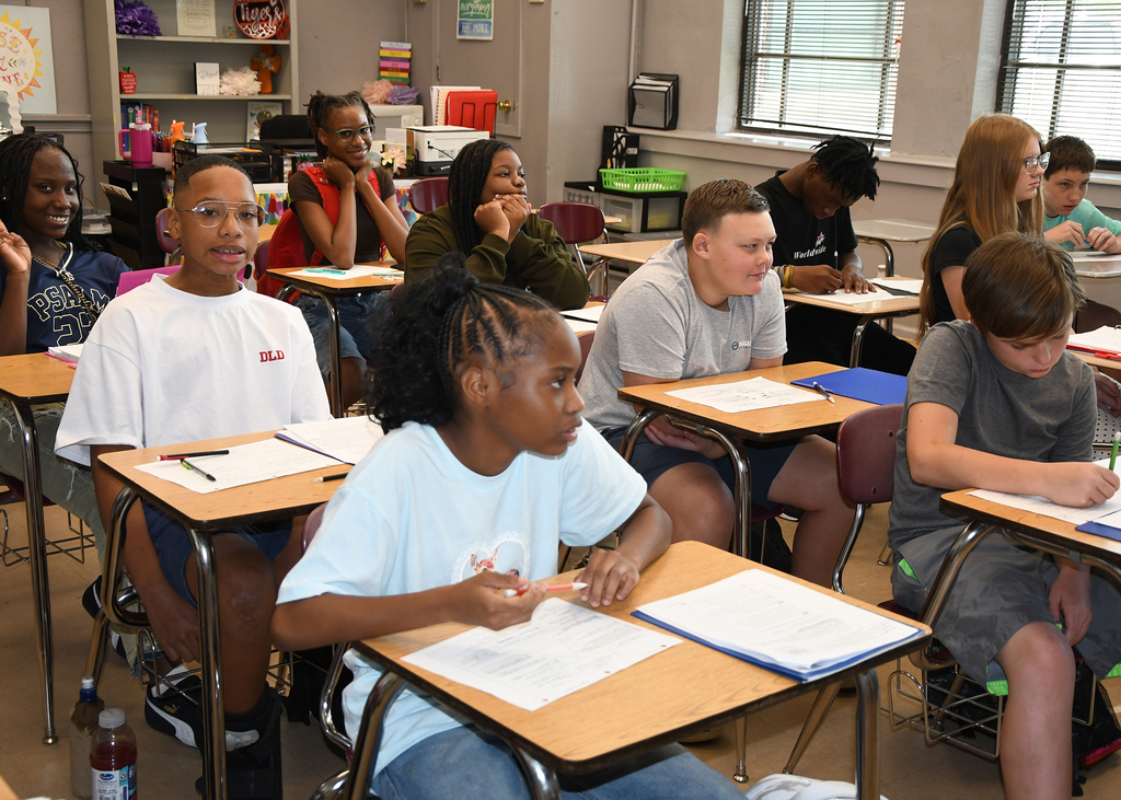 A classroom with students seated at desks, engaged in writing activities. Desks are equipped with papers, pencils, and books.