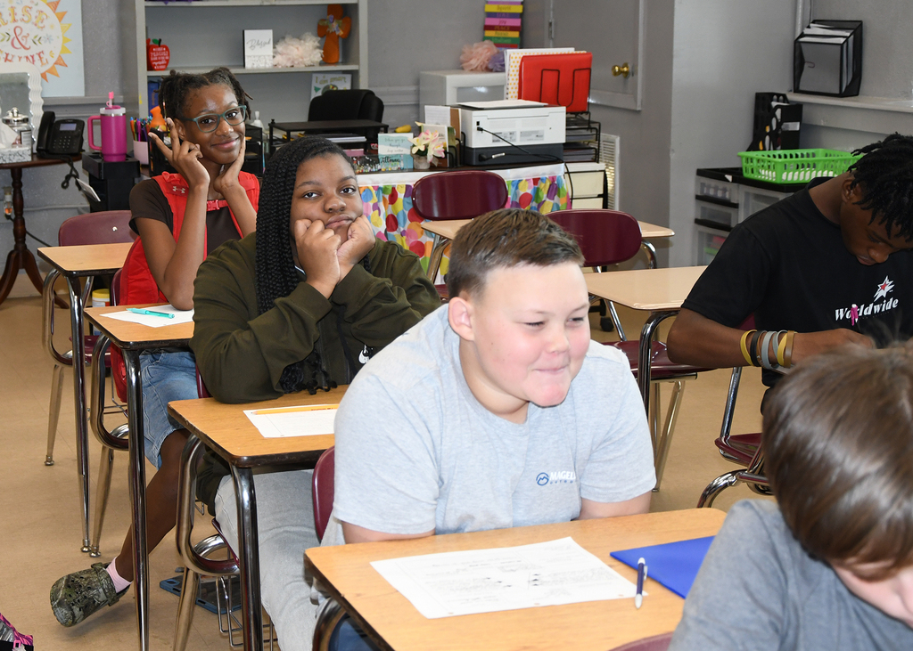 Students in a classroom, seated at desks, engaged in conversation, with one person talking on a phone.
