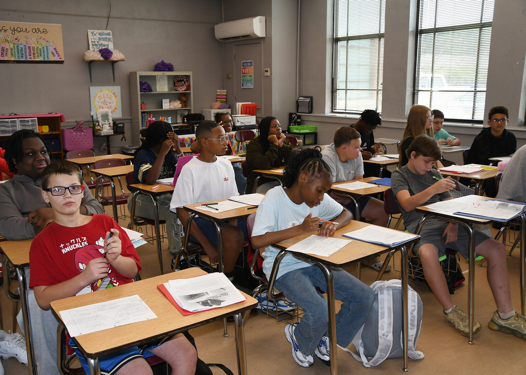 A classroom with students seated at desks, one wearing glasses, engaged in writing. Windows and posters adorn the walls.