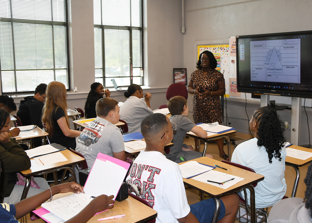 A classroom with desks and students. A teacher stands in front of a monitor displaying a graph.