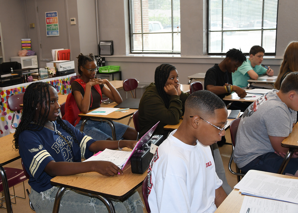 A classroom with students sitting at desks, working on laptops and papers. Desks have papers and laptops.