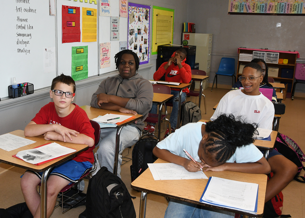 Students seated in classroom desks, working on assignments. Wall displays with various posters and bulletin boards.