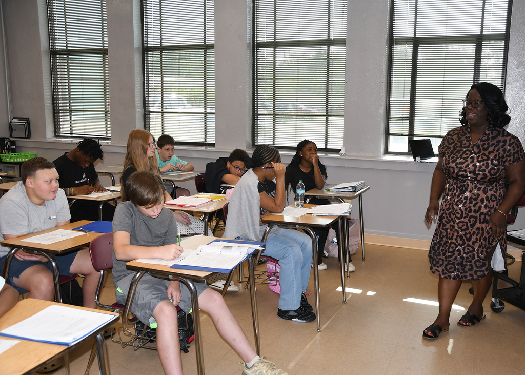 A teacher stands near a row of students seated at desks in a classroom with windows.