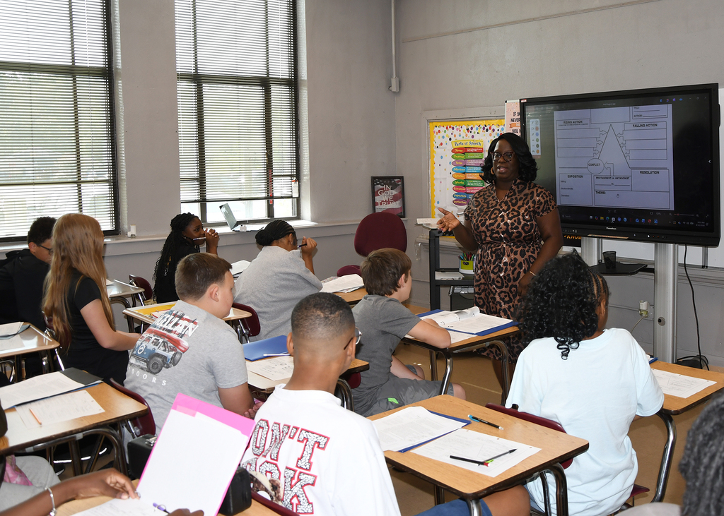 An instructor stands at a whiteboard, addressing students seated in a classroom with large windows and a monitor.