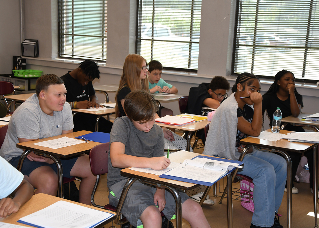 Students sit at desks in a classroom with large windows. Some students write while others look at their papers.