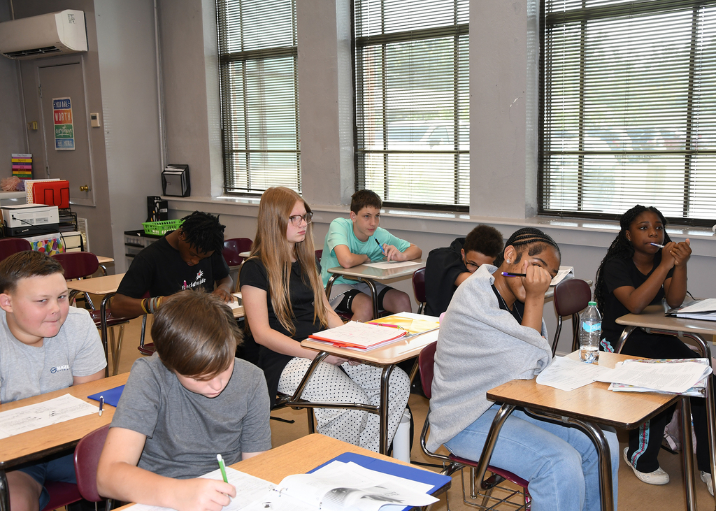 A classroom with students seated at desks. Some wear hoodies and sneakers, others wear t-shirts and jeans. One student uses a pencil to write.