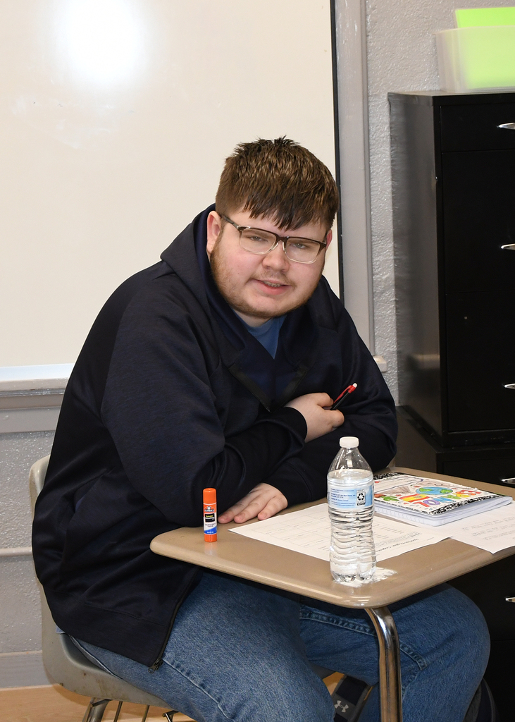 Person wearing glasses and a jacket seated at a desk with a pen, bottle of water, and books.