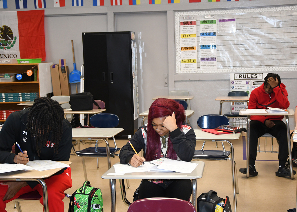 A classroom with several students seated at desks. Three students are seen working on papers.