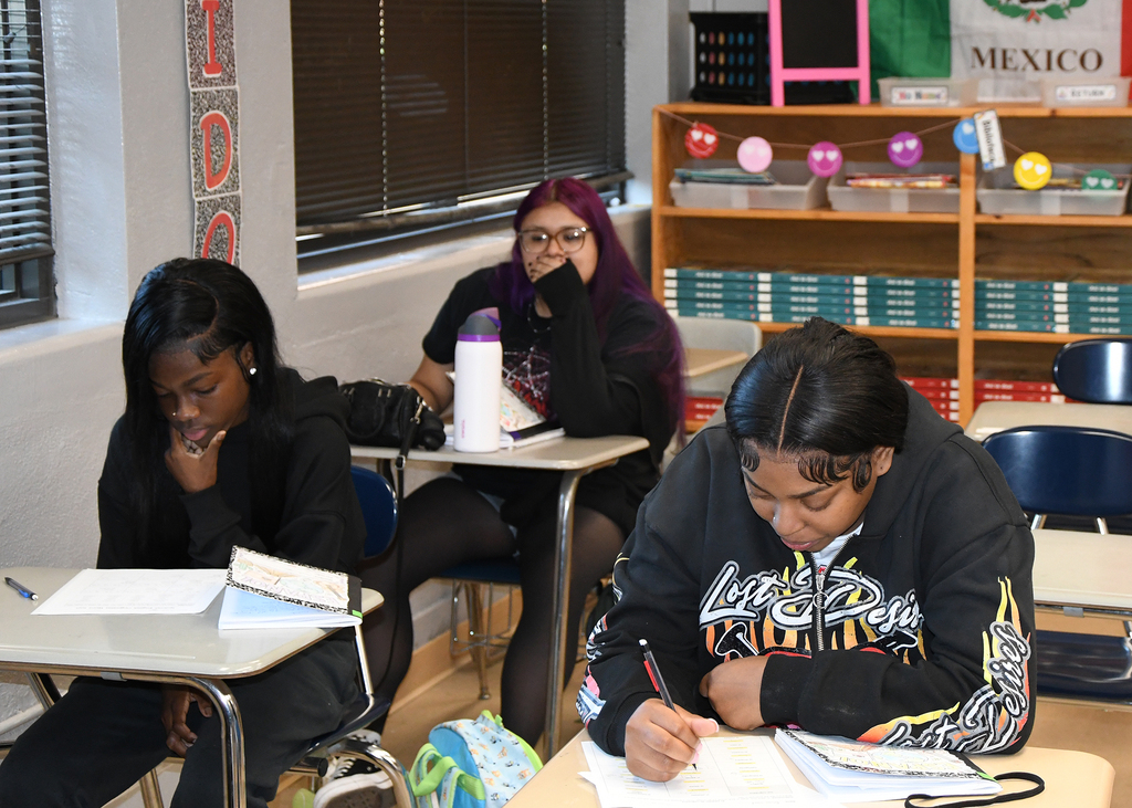 Three students seated at desks in a classroom, writing. One holds a pen, another has a book, and the third holds a water bottle.