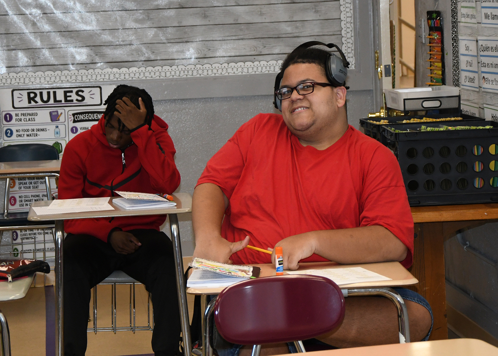 Two students in a classroom; one wears headphones and smiles while the other sits with hands covering face.