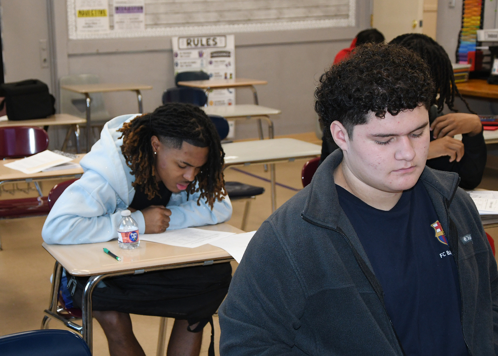Two students in a classroom, one writing while looking down, the other focused on their work.