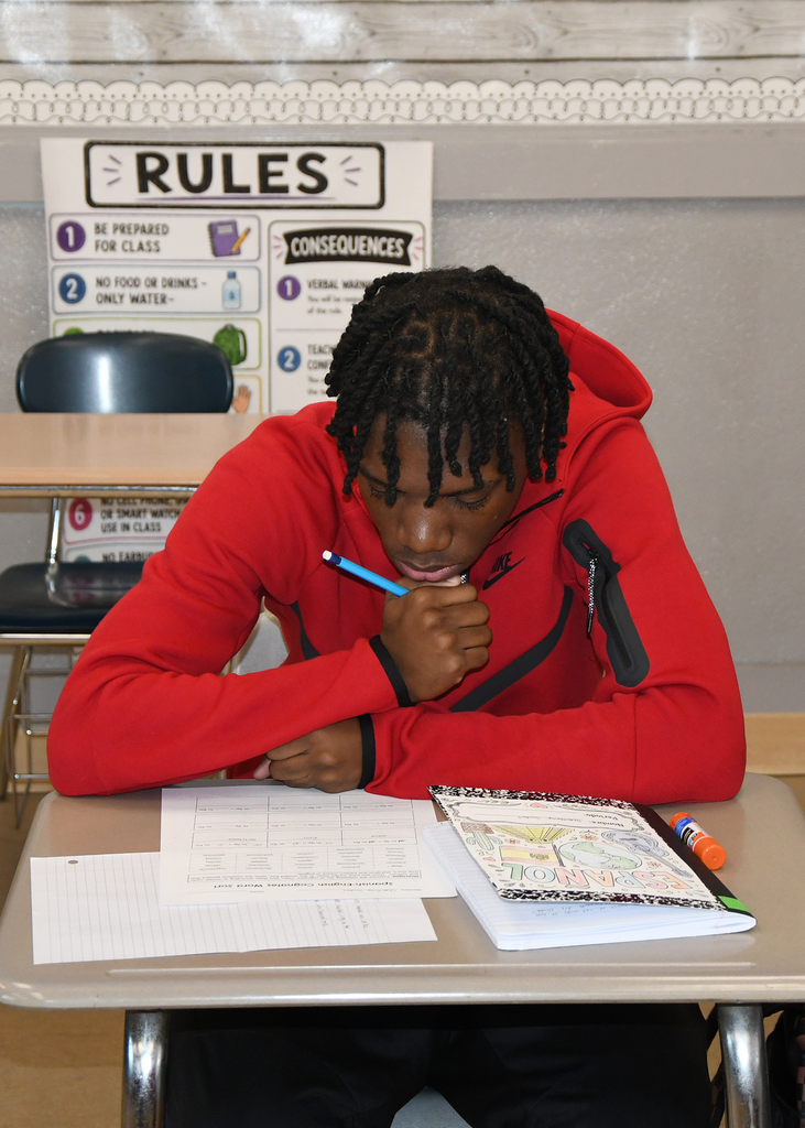 Person in red hoodie, seated at a desk, writing on a sheet of paper with a pencil.