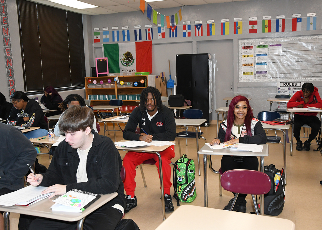 Students are seated at desks in a classroom with flags on the wall. One person in red pants is writing.