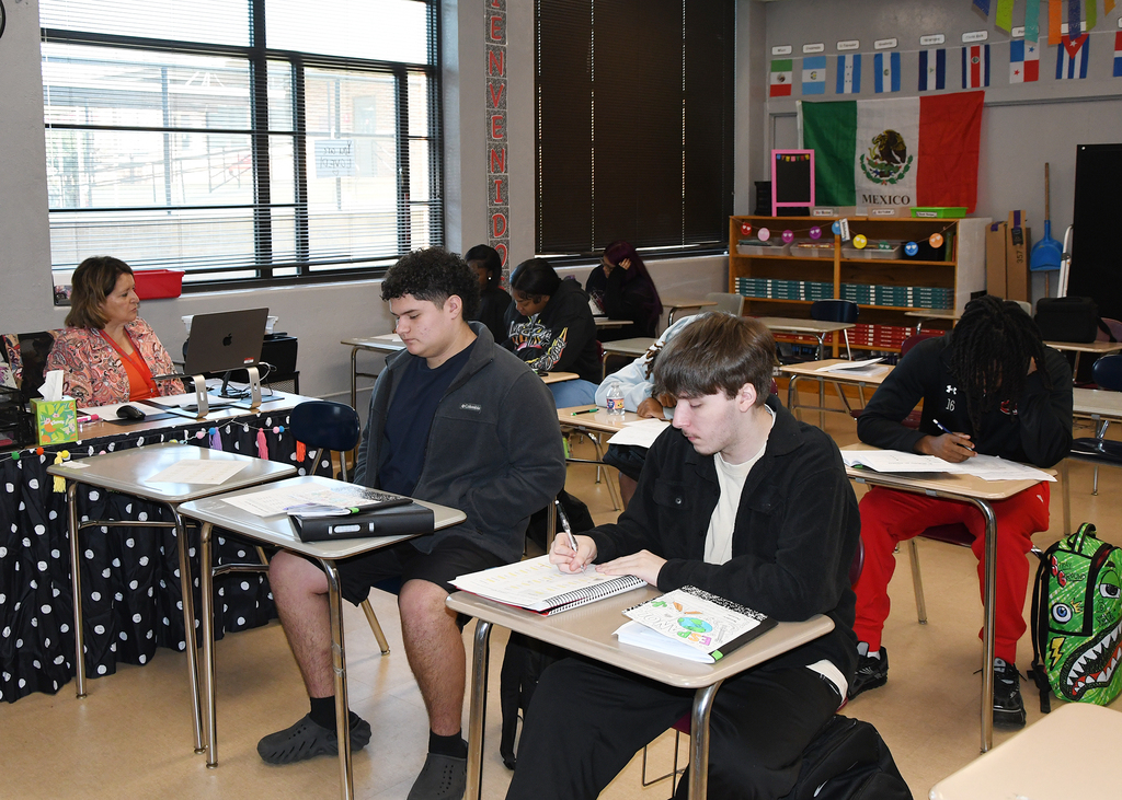 A classroom with desks and chairs. Students seated at desks, writing. One student has a backpack.