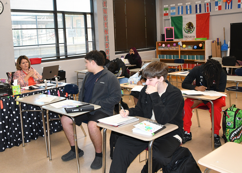 Students in a classroom, some sitting at desks, with a teacher at a table, a clock on the wall, and a Mexican flag.