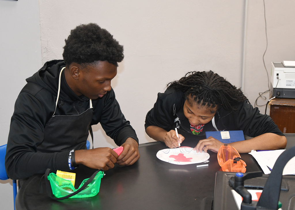 Two individuals in black hoodies and aprons, seated around a table, engaged in an activity involving a paper plate.