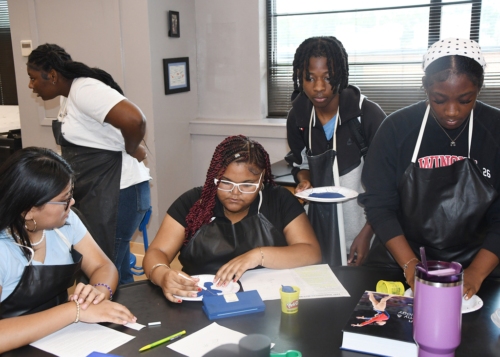 Five women in an art studio with aprons and glasses. One woman holds a paper with designs while another holds a purple cup.