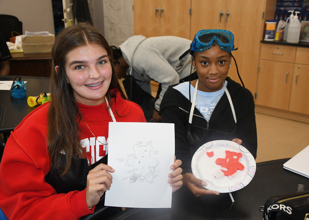 Two young people smiling, one holds a drawing, the other holds a red paper plate. Behind them, a cabinet with various items.