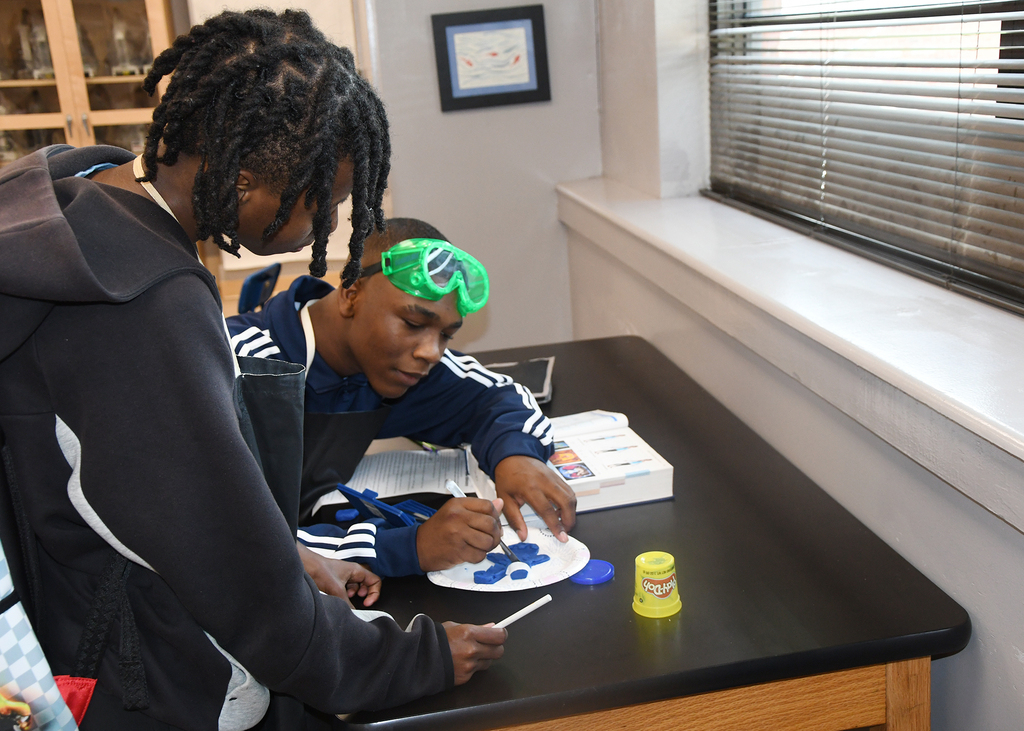 Two people at a table. One person is writing on paper while another observes. A picture is on the wall.