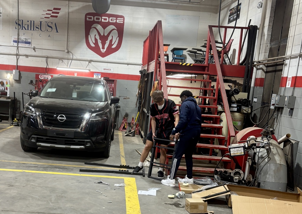 Two people work on a car inside a garage with a Dodge logo, stairs, and equipment.