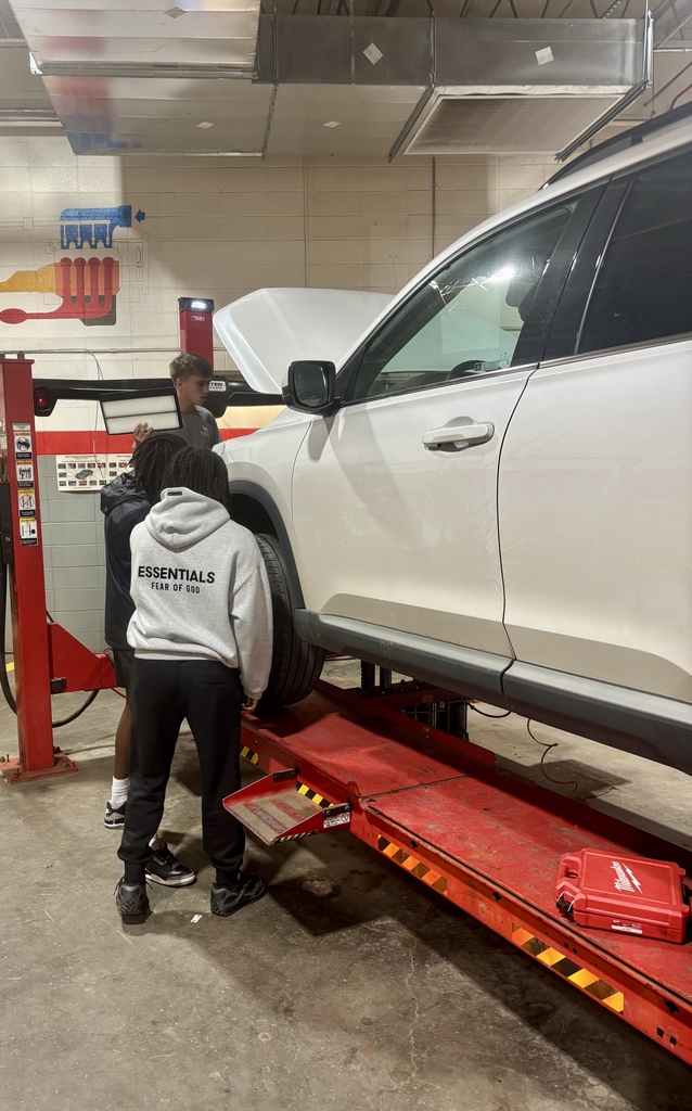Two people work on a white SUV with its hood open in a garage. One stands on a red lift platform.