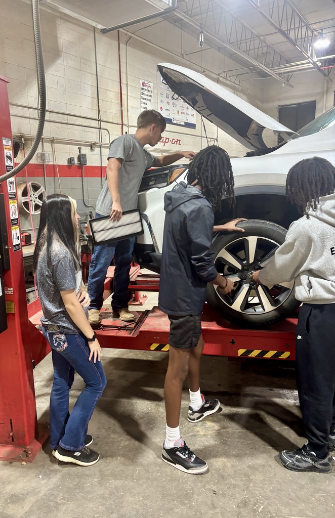 Four people work on a vehicle in a garage. Two men hold a wheel, one holds a tool, and a woman watches.