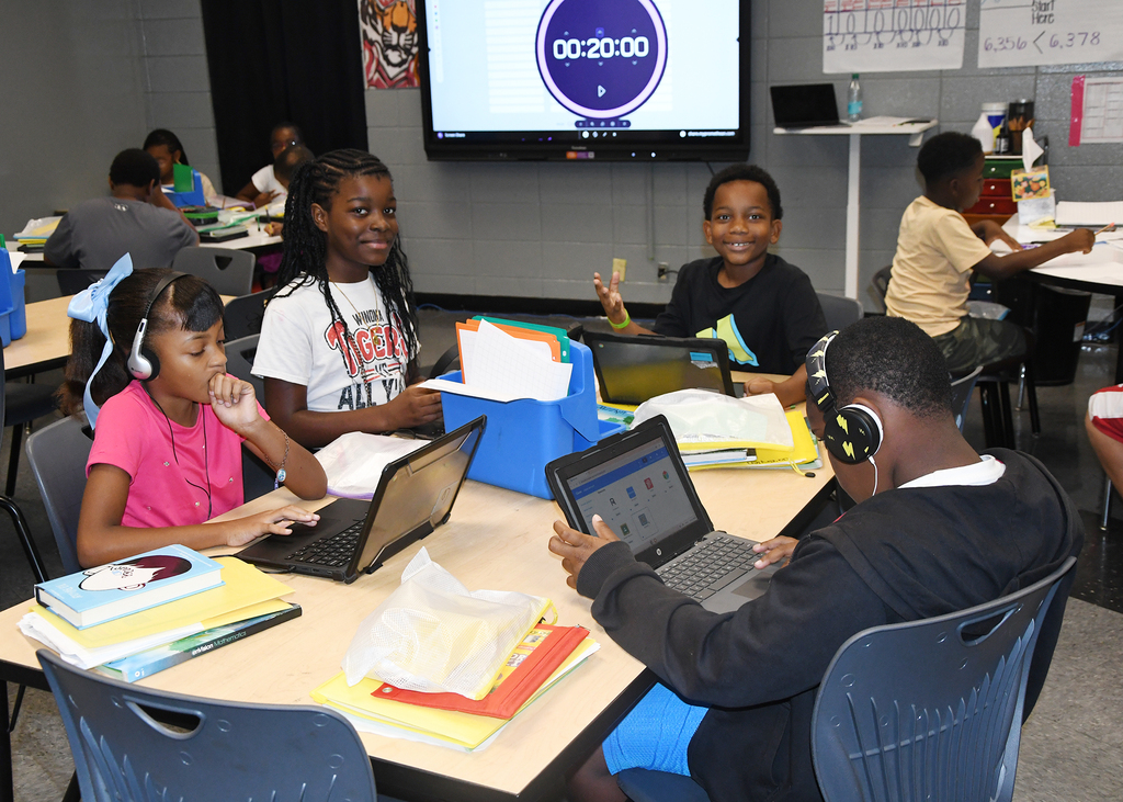 A classroom with students seated at tables. Some are using laptops, one has headphones on. A large monitor displays text.