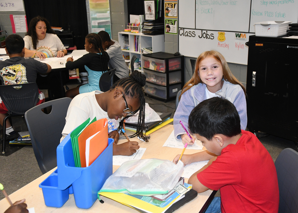 A classroom with children working at desks. A girl with red hair smiles. Two children write in notebooks.