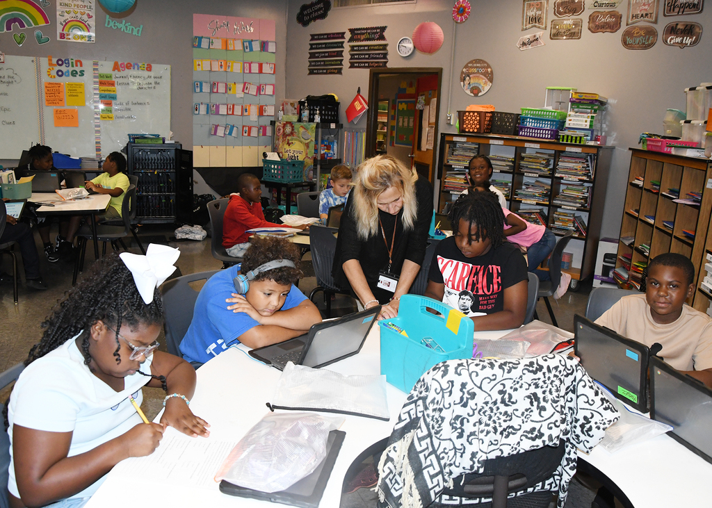 A classroom with students seated around a table, laptops, and a teacher in the middle.