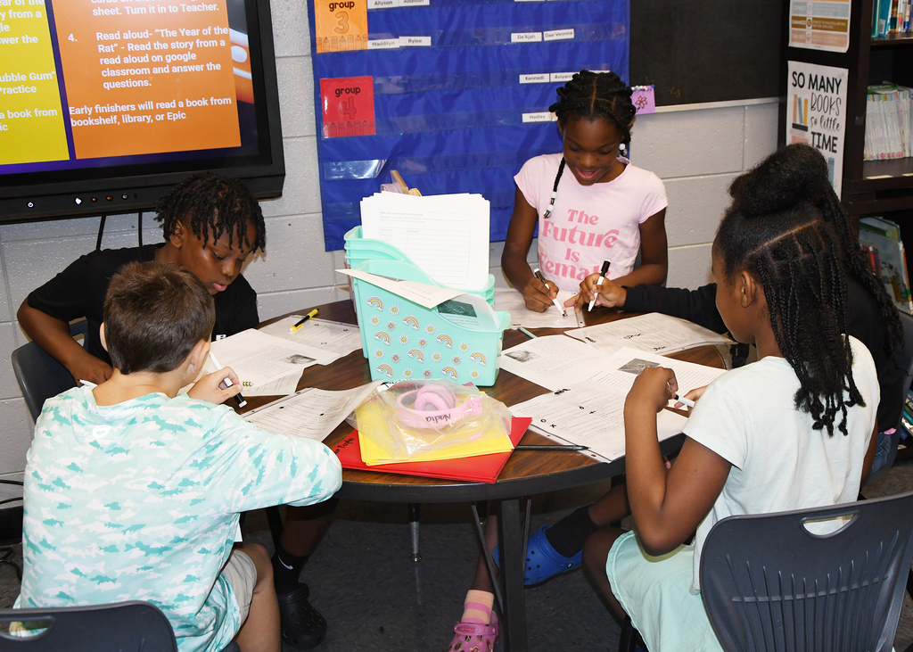 Five children sitting at a round table, engaged in writing activities. Background includes a TV, posters, and shelves with books.
