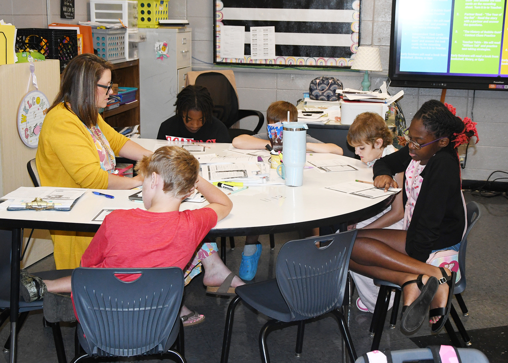 A classroom with a teacher and five children seated around a table. One child is using a marker.