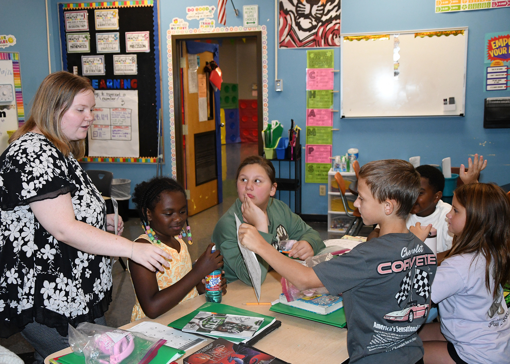 Classroom with blue walls. A woman and five students are seated around a table. One student holds a toothbrush.