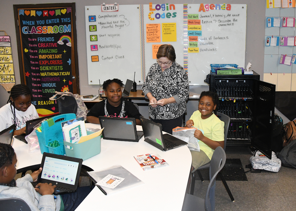 A group of kids are working on laptops at a table. A teacher is assisting them.